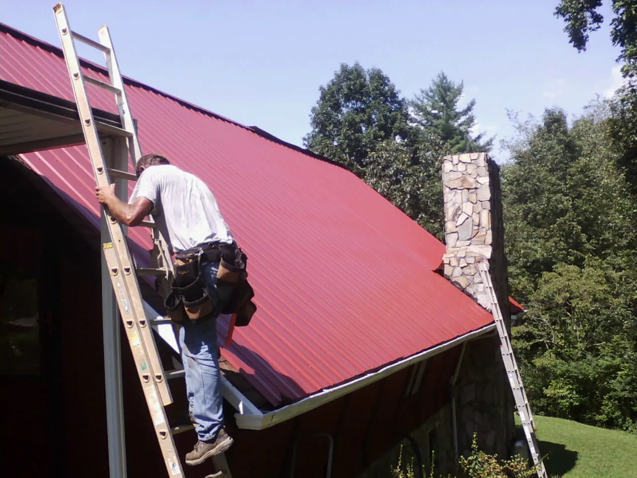 Roofer on a ladder beside a red metal roof and stone chimney — Soffit & Fascia Repair by Roofing Brothers in Hickory